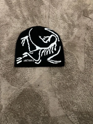 Svart mössa från Arc't - Säljer en svart mössa från Arc'teryx med stor vit logga i form av ett skelett på framsidan. Mössan är i mjukt material och har en clean, sportig look som passar perfekt till kalla dagar.
