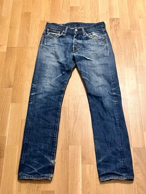 Levi's 501 blå jeans straight fit - Klassiska blå Levi's 501 jeans med rak passform och knappgylf. Jeansen har fem fickor och tydliga slitningar för en cool, avslappnad look. Perfekta för dig som gillar tidlös denimstil. W28 L32