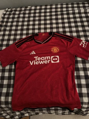 Manchester United matchtröja Adidas - Säljer en röd Manchester United matchtröja från Adidas med klubbmärke på bröstet och TeamViewer sponsortryck. Tröjan har svarta detaljer på axlarna och vita inslag vid kragen. Perfekt för fotbollsfans som vill bära sitt lag med stil.