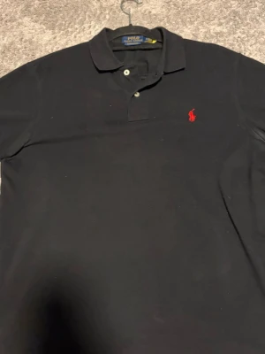 Svart pikétröja från Polo Ralph Lauren - Klassisk svart pikétröja från Polo Ralph Lauren i custom slim fit. Tröjan har krage, två knappar framtill och den ikoniska röda loggan broderad på bröstet. Perfekt till jeans eller chinos för en clean och stilren look.