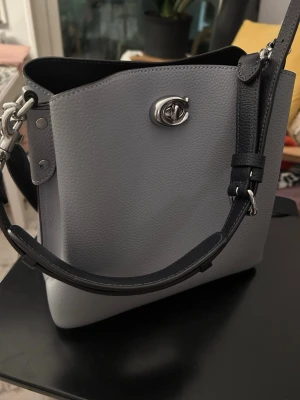 Grå handväska i skinn från Coach - Snygg grå axelväska från Coach i äkta skinn med silverfärgade metalldetaljer. Väskan har justerbar axelrem och smart vridlås framtill. Perfekt storlek för vardagens essentials och stilren design som passar till det mesta💙💙