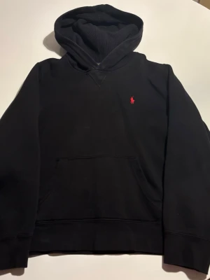 Svart hoodie från Polo Ralph Lauren - Svart hoodie från Polo Ralph Lauren med klassisk luva och känguruficka. Liten röd broderad logga på bröstet. Mjuk bomullskänsla och ribbade muddar vid ärmslut och nederkant. Perfekt för en avslappnad och stilren look.