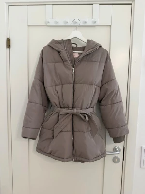 Beige pufferjacka med huva och bälte - Snygg beige pufferjacka med huva och bälte i midjan. Jackan har quiltad design, dragkedja framtill och långa ärmar. Perfekt för kyliga dagar och ger en trendig siluett. Passar 32-34