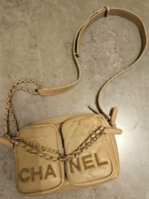 Beige axelväska från C h A n E l  - Säljer en beige axelväska från C h A n E l med två framfickor och kedjedetaljer på axelremmen. Väskan har en quiltad design på ena fickan och stora bokstäver med nitar som stavar märket bamr. Perfekt för dig som gillar lyxiga accessoarer.