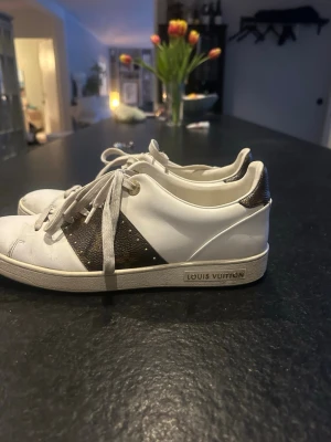 Louis Vuitton vita sneakers - Snygga vita sneakers från Louis Vuitton med klassiska bruna monogramdetaljer på sidan och hälen. Har använts ganska mycket och därav priset! Kan alltid diskutera priset och skriv för fler bilder! ❣️
