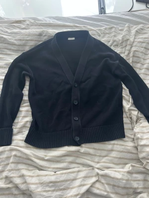 Svart kofta från John henric - Klassisk svart kofta från john henric med fyra knappar framtill och ribbade muddar vid ärmslut och nederkant. Koftan har en djup V-ringning och är tillverkad i mjukt stickat material, perfekt för lager på lager och enkel att matcha.