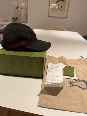 Svart Gucci keps med GG-mönster - Säljer en äkta Gucci keps i svart GG-mönstrad canvas med klassisk grön-röd detalj på sidan. Tidlös modell som passar till allt. 100% äkta finns kvitto   Detaljer: 	•	Märke: Gucci 	•	Färg: Svart 	•	Justerbar baktill 	•	Tillverkad i Italien 	•	Mycket fint skick  Kommer med: 	•	Originalbox 	•	Dustbag 	•	Kvitto & tags (se bilder)  Hör av dig vid frågor! 