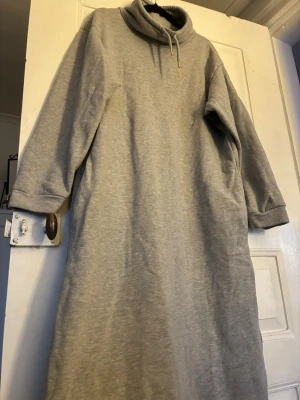 Grå sweatshirtklänning med polokrage - Mysig grå sweatshirtklänning från Holly & Whyte med polokrage och dragsko. Klänningen har långa ärmar och en avslappnad passform, perfekt för chill dagar hemma. Tillverkad i mjuk bomullsmix för extra komfort.