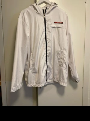 Vit vindjacka - Snygg vit vindjacka  med svart dragkedja framtill, huva och två sidofickor. Jackan har en bröstficka med röd logga och minimalistisk design. Perfekt för dig som gillar stilrena plagg med exklusiv känsla. Minus är att ena knappen på armen har gått av, men enkelt fixat för den händige. Köpt här och är osäker på om den är äkta därav priset.