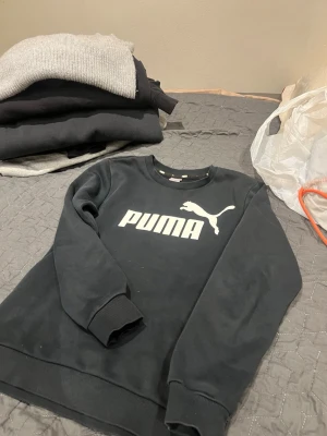 Svart sweatshirt från Puma - Snygg svart sweatshirt från Puma med stort vitt logotryck på bröstet. Tröjan har rund halsringning, ribbade muddar vid ärmslut och nederkant samt en klassisk avslappnad passform. Perfekt för en sportig och casual stil.