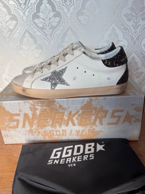 Golden Goose Superstar sneakers glitter - Golden Goose Superstar sneakers i vitt skinn med grå snörning, silverglittrig stjärna på sidan och glittrig häl i svart. Beige sula och tåparti i mocka. Klassisk låg modell med cool vintagekänsla och unika detaljer.