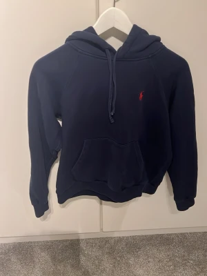 Mörkblå hoodie från Polo Ralph Lauren - Mörkblå hoodie med huva och känguruficka från Polo Ralph Lauren. Klassisk design med röd broderad logga på bröstet. Perfekt för en chill och stilren look. Mjuk bomullskänsla och avslappnad passform.