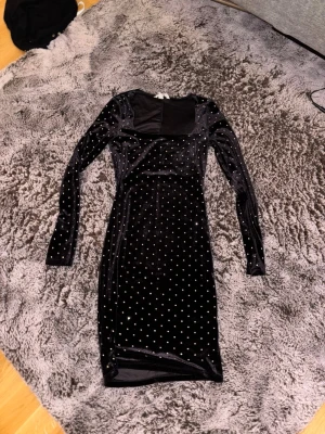 Svart sammetsklänning med paljetter - Svart långärmad sammetsklänning med fyrkantig ringning och glittriga paljettdetaljer över hela klänningen. Figurnära modell som passar perfekt när du vill sticka ut lite extra.