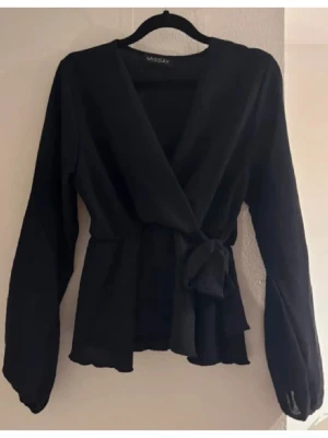 Omlottblus Missay - Stilren svart omlottblus från Missay med knytband i midjan och v-ringad hals. Blusen har långa ärmar och lätt utsvängd nederkant för en feminin siluett. Perfekt att matcha med både jeans och kjol.