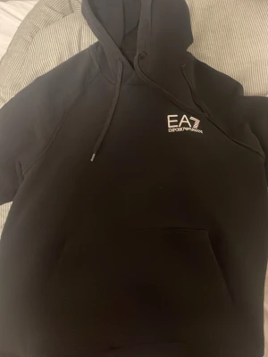 Svart EA7 Emporio Armani hoodie - Svart hoodie från EA7 Emporio Armani med vit logga på bröstet och stort tryck längs ena ärmen. Klassisk känguruficka framtill, huva med dragsko och ribbade muddar. Perfekt för en stilren och sportig look.