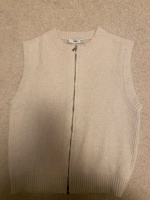 Beige stickad väst med dragkedja MNG - Beige stickad väst från MNG i storlek S. Västen har en rak passform, ribbade kanter och dragkedja framtill. Materialet är mjukt och ger en clean look som funkar till många olika stilar.