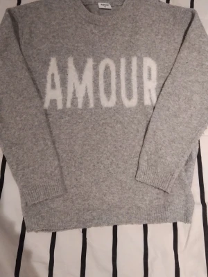Grå stickad tröja med text från Kappal - Mysig grå stickad tröja från Kaporal med stor vit text 'AMOUR' framtill. Tröjan har rund halsringning, långa ärmar och ribbade muddar vid ärmslut och nederkant. Perfekt för dig som gillar enkel men snygg design.💗