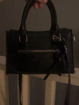 Svart handväska med dragkedja så snygg till alla tillfällen  - Snygg svart handväska i skinnimitation med både handtag och axelrem. Väskan har silverfärgade detaljer och en dragkedja framtill som ger en cool touch. Perfekt storlek för det viktigaste och passar till många olika outfits.