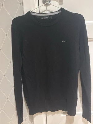 Svart tröja från J.Lindeberg - Stilren svart långärmad tröja från J.Lindeberg med rund halsringning och diskret logga på bröstet. Perfekt basplagg i garderoben, enkel att matcha och passar till många olika stilar.