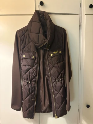 Brun barbour jacka  - Snygg brun quiltad dunjacka från Barbour med hög krage, gulddetaljer och två bröstfickor med knappar. Jackan har långa ärmar och dragkedja framtill. Perfekt för kyliga dagar och ger en stilren look.