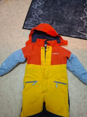 Overall från Didriksons - Säljer en färgglad täckbyxa från Didriksons med huva. Overallen har orange upptill, gula ben och blå detaljer på ärmar och ben. Utrustad med dragkedjor och reflexdetaljer. Perfekt för kalla vinterdagar och utomhuslek.