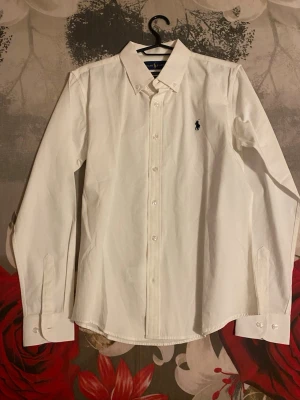 Vit skjorta från Ralph Lauren - Klassisk vit skjorta från Ralph Lauren med broderad logga på bröstet. Skjortan har lång ärm, knappar framtill och button-down krage. Tillverkad i mjuk bomull för en clean och tidlös look.