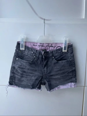 Svarta jeansshorts - One Green Elephant  - Snygga jeansshorts i svart med ljusrosa detaljer från One Green Elephant. Fransad kant, klassisk femficksmodell och tryckt logga i midjan. Storlek XS. Perfekta för sommarens festivaler, vardagsstil eller streetwear-look. Fint skick!