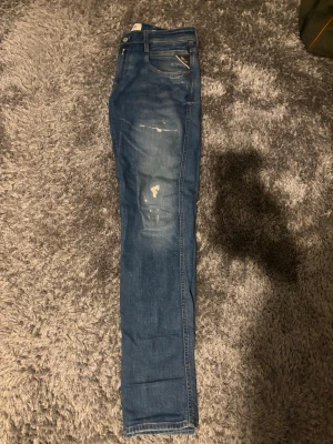 Blå replay jeans med slitningar - Säljer ett par blå replay anbass med slitna detaljer och tvättade partier. Jeansen har klassisk femficksdesign och smal passform. Perfekta för dig som gillar en avslappnad men trendig stil.