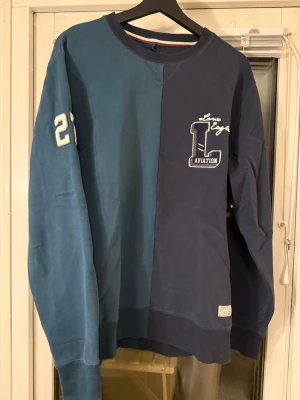 Tvåfärgad Lindbergh sweatshirt - Sweatshirt från Lindbergh i två nyanser, marinblå och grön, med Las Vegas Aviation-tryck och broderad siffra 21 på ärmen. Tröjan har rund halsringning, ribbade muddar och en unik delad design framtill och baktill. 