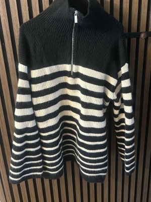 Svartvit randig stickad tröja med dragkedja - Snygg stickad tröja i svart och vitt med breda horisontella ränder. Tröjan har hög krage och halv dragkedja framtill. Långa ärmar och ribbstickad struktur upptill. Perfekt för kyliga dagar och enkel att matcha.