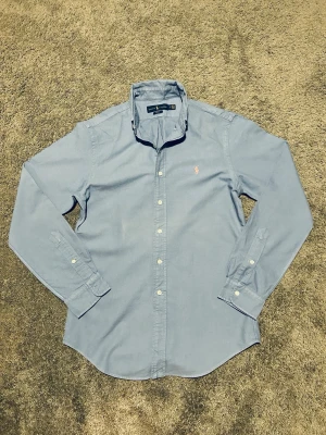 Blå skjorta från Ralph Lauren - Klassisk ljusblå skjorta från Ralph Lauren med broderad logga på bröstet. Skjortan har knappar framtill och vid ärmsluten samt en stilren krage. Perfekt för en clean och tidlös look.