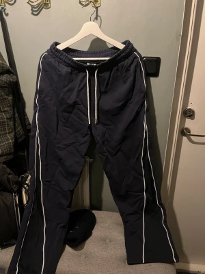 Mörkblå mjukisbyxor med vita ränder Gymshark  - Mörkblå mjukisbyxor från Gymshark med vita ränder längs benen och bred resår i midjan. Byxorna har snörning framtill och raka ben. Perfekta för en avslappnad stil.