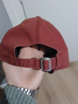 Vinröd keps från New Era - Säljer en stilren vinröd keps från New Era med justerbart metallspänne bak. Kepsen har klassisk form och är tillverkad i bomullstyg. Broderad New Era-logga på sidan ger en clean och sportig vibe.