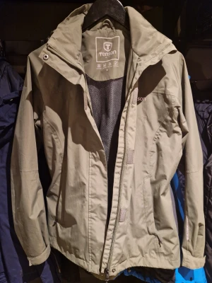 Knappt använd!Supersnygg grön regnjacka från Tenson - Stilren olivgrön regnjacka från Tenson med huva och dragkedja framtill. Jackan har en enkel design, meshfoder på insidan och justerbar huva. Perfekt för blåsiga dagar och outdoor-äventyr. Klassisk passform och helt vattentät. Knappt använd, nyskick!