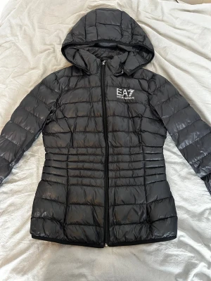 Svart dunjacka från EA7 Emporio Armani - Snygg svart dunjacka från EA7 Emporio Armani med huva och quiltad design. Jackan har dragkedja framtill och logga på bröstet. Perfekt för kalla dagar och ger en sportig, modern look.