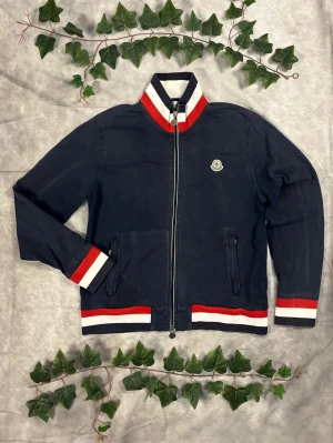 Moncler Manglia Cardigan – Navy/Tricolour – Strl L - |Färg: Navy med röd/vit/blå ribb, tydligt blekt | Modell: Manglia Cardigan, zip | Storlek: L, normal–slim passform | Skick: Grade C, 6,5/10 (missfärgad) | Hör av dig ifall du har funderingar!
