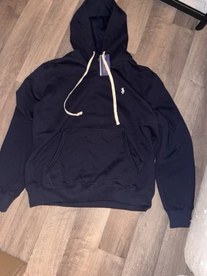Mörkblå hoodie från Polo Ralph Lauren - Mörkblå hoodie med vit dragsko och klassisk Polo Ralph Lauren-logga broderad på bröstet. Hoodien har en stor känguruficka framtill och ribbade muddar vid ärmslut och nederkant. Perfekt för en chill och stilren look.