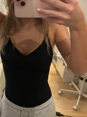 Svart body med smala axelband - Snygg svart body med smala axelband och v-ringning. Modellen har en lätt rynkning vid bysten och är tillverkad i ett stretchigt, mjukt material som sitter tight mot kroppen. Bodyn har även knäppe.💗