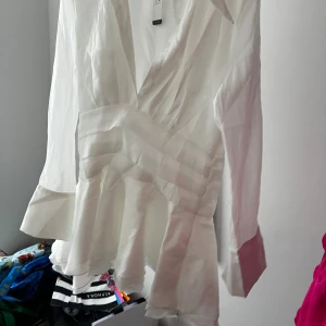 Vit volang klänning - Säljer en vir volangkkänning med v-ringning, markerad midja och volangdetaljer nedtill. Blusen har långa ärmar och är tillverkad i ett lätt, glansigt material som ger en lyxig känsla. Aldrig använd lapp kvar