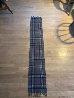  halsduk från Amanda Christensen - Klassisk grå halsduk med rutigt mönster i svart, vitt och röda detaljer. Halsduken har fransar i ändarna och är tillverkad i mjuk kashmir som håller dig varm. Perfekt accessoar för att lyfta din outfit under kyliga dagar.