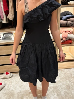 Svart oneshoulder klänning med volang - Snygg svart kortklänning med oneshoulder-design och stor volang över axeln. Kjolen är lätt utställd och har en skrynklig look. Perfekt för dig som vill sticka ut med en unik och trendig stil. Man kan snurra i den riktigt bra! Pris kan diskuteras 