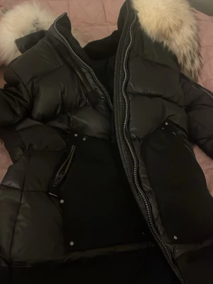 Svart dunjacka Rockandblue med pälskrage - Säljer en svart dunjacka från Rockandblue med stor fluffig pälskrage och flera dragkedjor. Jackan har quiltad design, lång ärm och snygga detaljer som logga på ärmen. Perfekt för kalla dagar och riktigt trendig look.