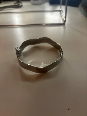 Silvrigt vågformat armband - Ett stilrent armband i silverfärgad metall med vågig design. Armbandet har en slät, blank yta och är stelt i modellen vilket ger en modern och minimalistisk look. Perfekt för dig som gillar unika accessoarer med enkel touch.