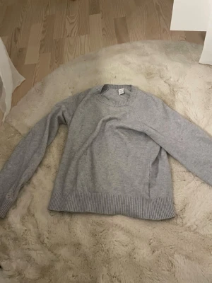 Ljusgrå stickad tröja H&M - En enkel och stilren ljusgrå stickad tröja från H&M. Tröjan har rund halsringning, långa ärmar och bred ribbstickad mudd vid ärmslut och nederkant. Perfekt basplagg för lager på lager och passar till det mesta.