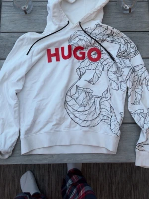 Vit hoodie från HUGO med tryck - Snygg vit hoodie från HUGO med stor röd logga på bröstet och svart/vitt blommönster över axel och ärm. Hoodien har huva med svarta snören och ribbade muddar. Perfekt för dig som vill sticka ut med en unik design. Pris går att diskutera!
