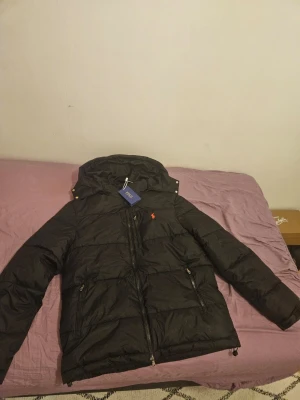 Polo rahlp lauren puffer - Snygg svart dunjacka från Polo Ralph Lauren med huva och dragkedja framtill. Jackan har två sidofickor med dragkedja och den klassiska röda loggan broderad på bröstet. Perfekt för kalla dagar och riktigt stilren look.