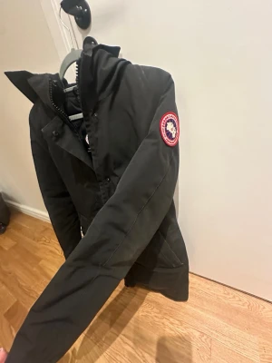 Svart parkas från Canada Goose - Säljer en svart parkas från Canada Goose med klassisk patch på ärmen och huva med spänne. Jackan har dragkedja och knappar framtill samt en rak passform. Perfekt för kalla dagar och har en stilren look. Använd men ingen synligt!