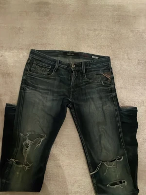 Replay Anbass blå distressed jeans - Säljer ett par Replay Anbass jeans i blå tvätt med slitningar på framsidan och distress på baksidan. Klassisk femficksmodell med normal passform och låg midja. Snyggt slitna för en cool och avslappnad look. Materialet är sopigt mjukt jeans.