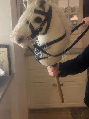 Käpphäst med vit man och träns - Säljer en käpphäst med mjukt vitt huvud, svart träns och tyglar. Käppen är i trä och hästen har broderade detaljer och realistiska ögon. Perfekt för lekar och fantasiäventyr. 500 för bara hästen, 550  för tyglarna. Tyglarna kostar 50. 