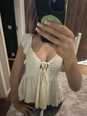 Vit omlottopp med knyt och volang - Supersöt vit topp med omlottdesign, knyt framtill och volangkant. Ärmlös med breda axelband och lätt rynkad struktur. Perfekt till jeans eller kjol för en fräsch look. Materialet känns som bomull eller linne.